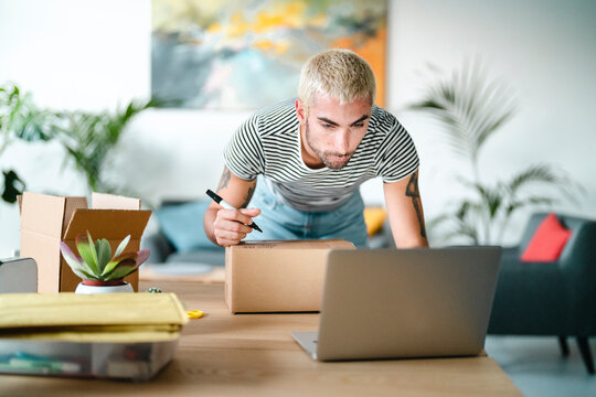 A young man with blonde hair writes on a cardboard box with a marker. He is preparing packages for shipping, managing his online business from home with a laptop nearby.