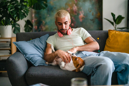 A young man with pink headphones sits on a sofa, affectionately petting his small dog. The dog is relaxed, lying on its back, enjoying the moment.