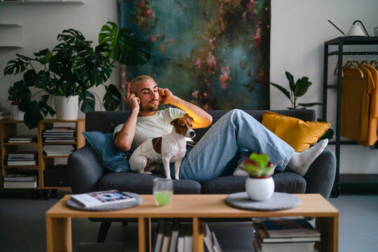 A man with blonde hair and a beard relaxes on a sofa, listening to music through headphones with his eyes closed. His small Jack Russell Terrier dog sits calmly beside him in a modern living room.