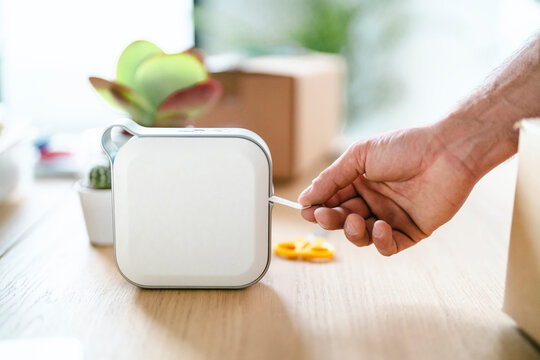 A man's hand pulls a freshly printed label from a sleek white label printer. The device rests on a light wooden desk, surrounded by office supplies, suggesting organization or shipping tasks.