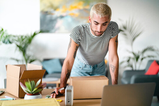 A young man with blonde hair and tattoos is preparing packages for shipment at home. He stands over a desk with cardboard boxes, a laptop, and a marker, focusing on his task.