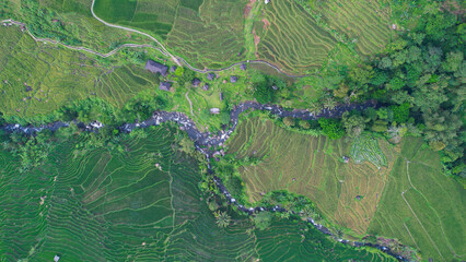 Aerial view of terraced paddy fields in Majalengka, Indonesia, showcasing vibrant green terraces, a winding river, and scattered huts amidst the verdant landscape.
