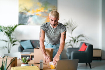 A young man with blonde hair and tattoos is standing at a table, carefully handling a wrapped item near a cardboard box. He is packing or unpacking items in a modern living room.