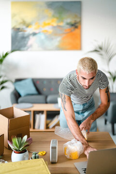 A young man with blonde hair and tattoos is carefully wrapping items in bubble wrap on a wooden table. He is packing boxes in a bright living room, possibly preparing for a move or shipping.
