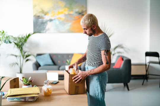 A man with blonde hair and a striped shirt packs an item into a cardboard box on a wooden table. He is preparing packages for shipment in a home office setting.