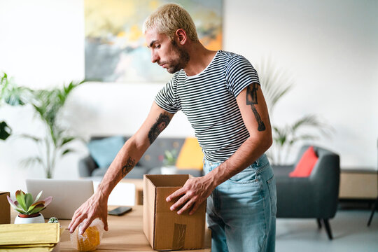 A young man with blonde hair and tattoos packs a bubble-wrapped item into a cardboard box on a wooden table. He is preparing a shipment or organizing belongings in a modern home.