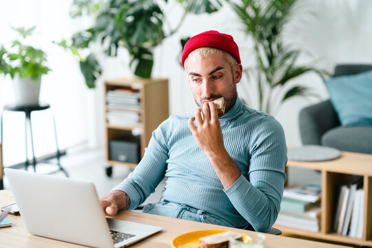 A man in a red beanie and blue shirt works on his laptop at a desk. He takes a bite of food from his hand while focusing on the screen. This scene depicts a casual work-from-home environment.