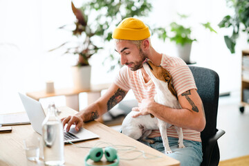 A man in a yellow beanie works on his laptop at a desk. His small dog sits on his lap, licking his face. This scene shows a relaxed home office environment with a pet.