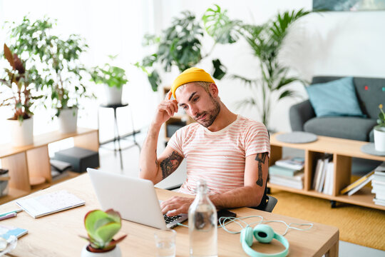 man with a yellow beanie and tattoos, wearing a striped shirt, works intently on his laptop at a modern wooden desk. He is deep in thought, surrounded by lush green plants in a home office.