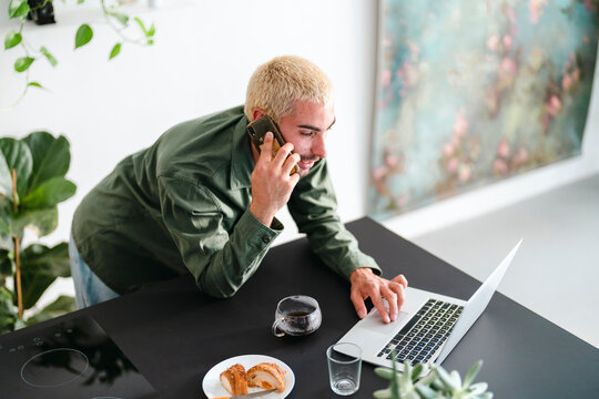 A man with blonde hair talks on his smartphone while working on a laptop at a modern kitchen counter. He has coffee and a croissant nearby.