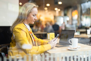 Business woman writes message on smartphone while sitting at table in a cafe. View through window