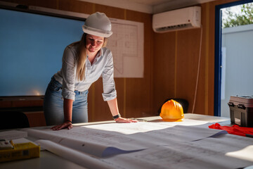 Female architect wearing a white hard hat leaning over blueprints spread out on a table inside a...