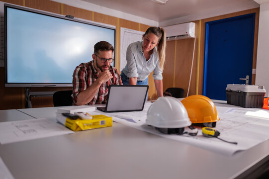 Two engineers are analyzing blueprints and working on a laptop in their office container, discussing the progress of their construction project - Powered by Adobe