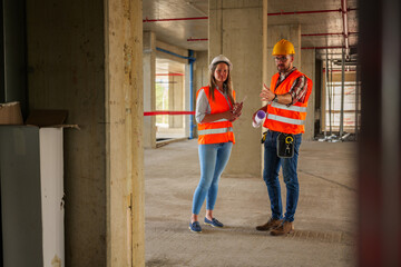 Two construction workers, a man and a woman wearing safety vests and helmets, are discussing a...