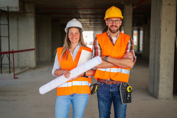 Two architects, a man and a woman, smiling while holding blueprints, wearing safety vests and...