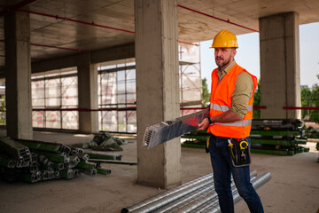 Construction worker wearing a safety vest and hardhat, carrying a heavy metal beam while diligently...