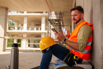 Construction worker taking a coffee break, sitting and relaxing while using a smartphone at a...