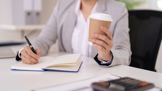 Dedicated professional adult hands writing important business notes and taking down ideas while holding a warm coffee cup at a modern office workspace desk fostering productivity