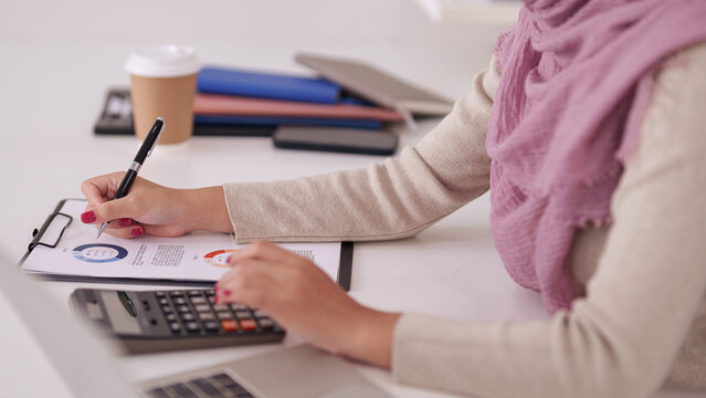 Asian adult Muslim businesswoman diligently analyzing financial data reports at her office desk using a pen and calculator for strategic business planning