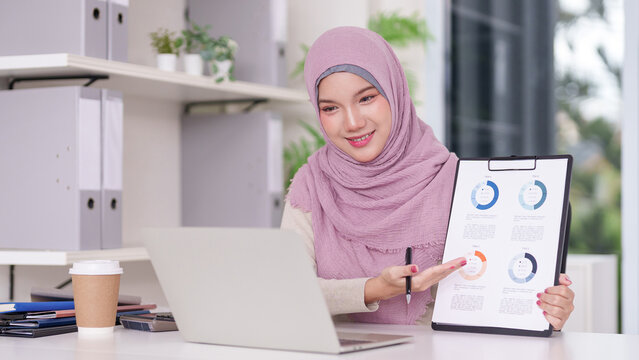 Asian adult Muslim businesswoman presenting financial data charts during online video conference for business growth and professional development in modern office environment - Powered by Adobe