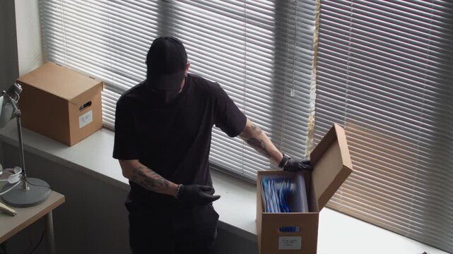 High angle shot of male criminal hacking into corporate computer in office while talking to partner rummaging through documents in box searching for confidential data during industrial espionage