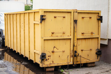 Rusty yellow dumpster waits silently in the alley behind a busy urban street on a cloudy day