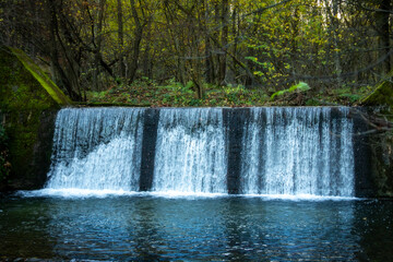 Fototapeta premium Small forest waterfall on a stone dam in autumn scenery, surrounded by mossy trees and serene reflections — peaceful nature escape in southern Poland woods