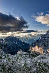 Obraz premium Rugged peaks and dramatic valleys in the Dolomites at sunset — vertical aerial view with scattered light on alpine ridges, dark forests below, and heavy clouds above the Italian Alps