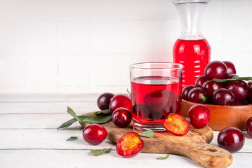 Sweet and tasty red cherry plum juice in in jug and glass, with fresh cherry plum fruits and tree leaves on a white background, copy space