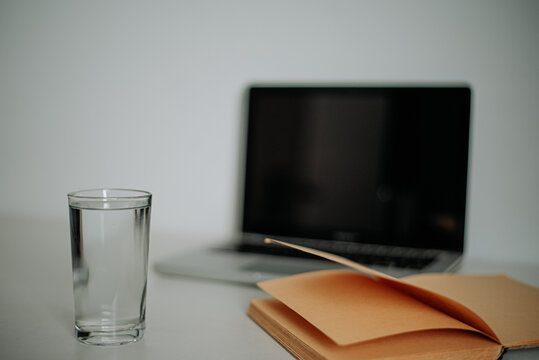 Glass of water, laptop and open notebook on clean white surface, calm work scene.