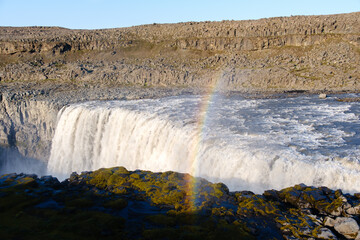 Majestic Dettifoss waterfall pours into a canyon, creating mist that catches sunlight and forms a...