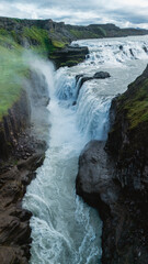 A stunning display of natures power as Gullfoss waterfall flows dramatically into a deep gorge. Thick mist rises from the rushing waters under a cloudy sky, showcasing Icelands wild beauty.