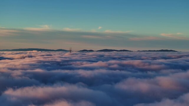 Aerial view of Sutro Tower poking through a sea of fluffy clouds, with mountains in the distance under a blue sky, San Francisco, California, United States.