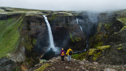 Two hikers stand at the edge of a dramatic cliff, gazing at the powerful Haifoss waterfall...