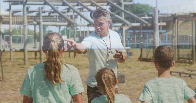 Pointing coach wearing whistle instructing children on sports field, with obstacle course - Powered by Adobe