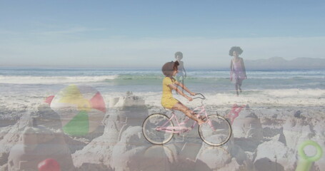 Pedaling girl in yellow dress riding pink bicycle on beach, with sandcastles, beach ball and shovel