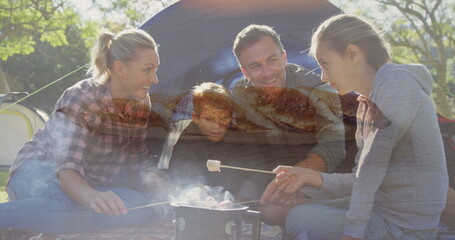 Family kneeling and sitting around camp stove roasting marshmallows in campsite, with dome tent