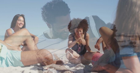 Laughing friends sitting on white sand at beach, wearing tank tops and swim trunks with sunglasses