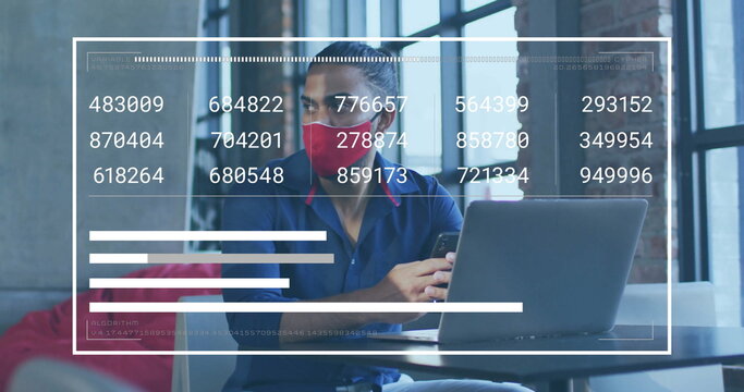 Analyzing overlaid data on laptop, man in red mask blue shirt holding smartphone at coworking desk