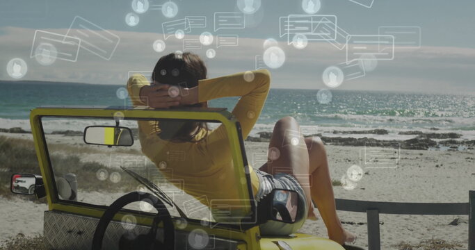 Leaning back woman wearing yellow top, striped shorts in dune buggy on shoreline, with chat icons - Powered by Adobe