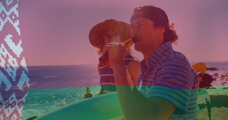 Standing two men in beachwear holding surfboards and sipping amber beer bottles on rocky beach