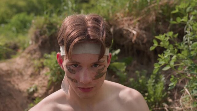 Brown hair tourist stares into camera under bright sunlight, mud paint smeared on cheeks, headband in place. Slow motion reveals calm yet bold expression, conveying silent command on rugged terrain.