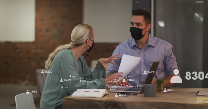 Masked colleagues reviewing papers at office desk, with laptop, plant and digital overlay