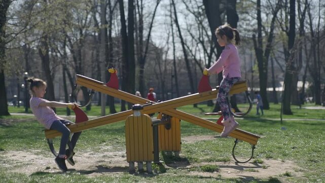 Little girls swing on the see-saw at the playground in the park, with trees in the blurred background, during the spring season.