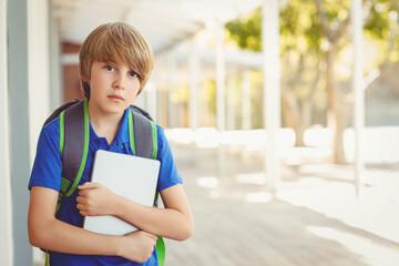 School-age boy holding tablet while standing on school walkway wearing blue polo shirt and backpack