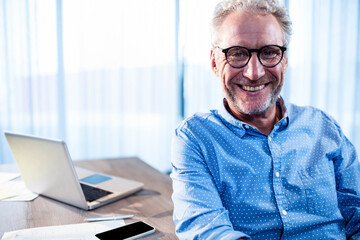 Senior man wearing glasses sitting at desk using laptop, phone, sorting papers by bright window