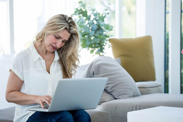 Woman typing on silver laptop while sitting on light-colored sofa under floor-to-ceiling windows