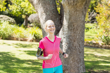 Woman jogging past oak tree through park in sportswear using phone armband with fitness watch