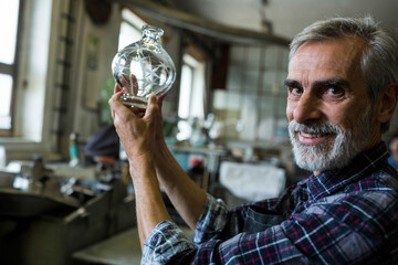 Senior craftsman holding cut glass vase up toward light in workshop with workbenches, copy space