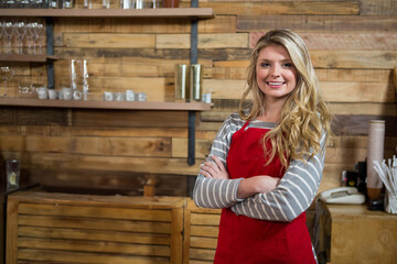 Female barista wearing red apron and shirt standing in cafe smiling with water glass, mugs behind